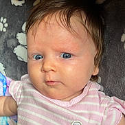 Liliana is registered to the contest to win money with this photo: baby, infant, child, blue_eyes, pink_clothing, striped_onesie, bear_patch, blanket, soft_texture, curious_expression, face, head, hair, skin, lying_down, indoors, portrait, closeup, cute, person