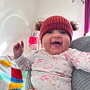 Shelsea a rejoint le concours — aidez-le/la à gagner de superbes lots ! baby, smiling, red_hat, knit_hat, pom_poms, floral_onesie, baby_clothing, happy, indoor, colorful_blanket, crochet_blanket, adult_hand, bracelet, nail_polish, air_conditioner, curtains, crib, person, child, portrait