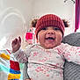 baby, smiling, red_hat, knit_hat, pom_poms, floral_onesie, baby_clothing, happy, indoor, colorful_blanket, crochet_blanket, adult_hand, bracelet, nail_polish, air_conditioner, curtains, crib, person, child, portrait