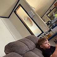 Michael is registered to the contest to win money with this photo: child, boy, couch, living_room, indoors, device, focused, casual, furniture, home, relaxed, glass_door, seated, short_hair, black_shirt, household_items, interior, young, technology, portrait