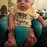 baby, background, blue_baby_seat, casual_clothing, child, expression, face, feet, furniture, hand, head, home, indoor, infant, legs, person, shirt, shoes, text_on_shirt, tongue_out