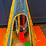 child, toddler, play_tent, toys, colorful, orange_mat, blue_cabinet, indoor, soft_rug, blocks, striped, yellow_tent, red_stars, curly_hair, playing, floor, striped_toy, striped_block, striped_ribbon, young_child