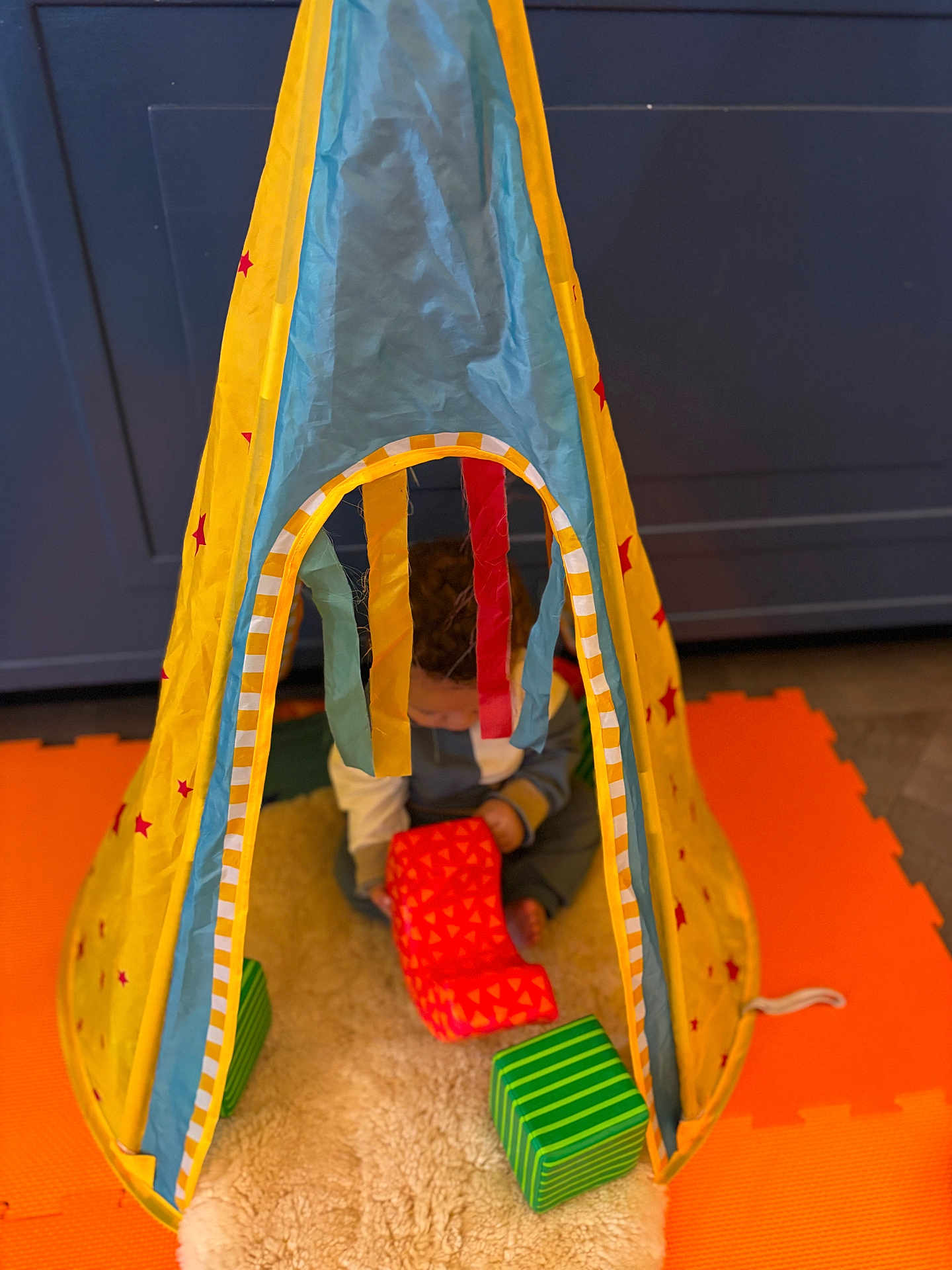 Cole is registered to the contest to win money with this photo: blocks, blue_cabinet, child, colorful, curly_hair, floor, indoor, orange_mat, play_tent, playing, red_stars, soft_rug, striped, striped_block, striped_ribbon, striped_toy, toddler, toys, yellow_tent, young_child