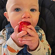 Theodore is registered to the contest to win money with this photo: baby, child, blue_eyes, strawberry, fruit, denim_overalls, long_sleeve_shirt, patterned_clothing, high_chair, close_up, cute, hand, face, eating, food, indoor, infant, person, skin, expression