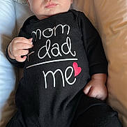 Benjamin Schneider a rejoint le concours — aidez-le/la à gagner de superbes lots ! baby, infant, onesie, black_clothing, text_on_clothing, heart_symbol, cushion, pillow, lying_down, indoors, child, portrait, cute, expression, person, skin, hand, face, head, soft_lighting