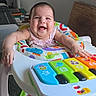 baby, baby_walker, toy_piano, colorful, smiling, happy, infant, indoor, child, playtime, musical_toy, seat, foot, arms, face, clothing, furniture, joy, expression, home