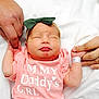 Amy is registered to the contest to win money with this photo: adult_hands, baby, closeup, cute, family, green_bow, headband, holding_fingers, hospital_id_band, infant, newborn, nursery, parental_care, pink_clothing, portrait, ring, skin, sleeping_face, tongue_out, white_sheet