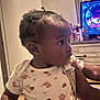 Gerzi is registered to the contest to win money with this photo: baby, child, indoor, onesie, patterned_clothing, curious, hands, television, furniture, person, hair_ties, infant, holding, cute, side_profile, young_child, room, floor, skin, portrait