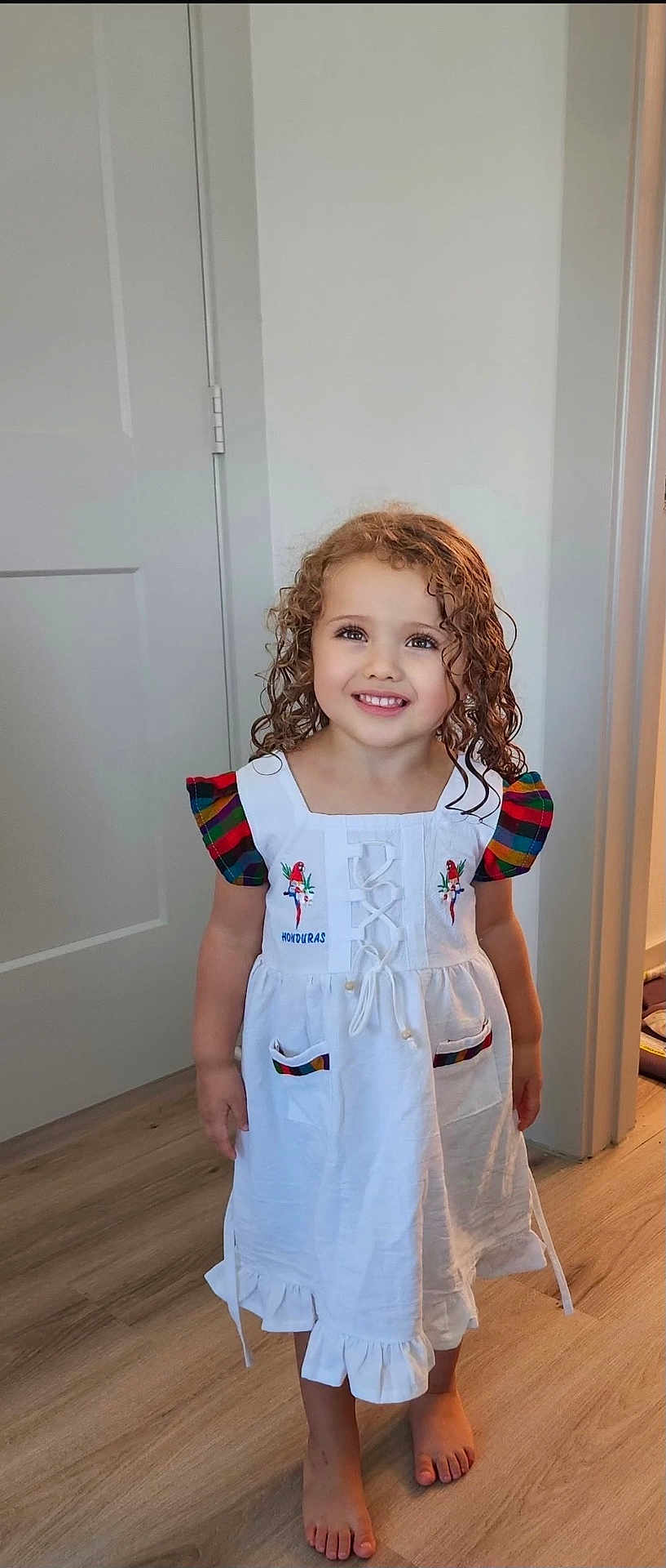 Gianna is registered to the contest to win money with this photo: child, girl, curly_hair, smiling, white_dress, barefoot, indoor, wooden_floor, colorful_sleeves, embroidery, parrot, happy, portrait, standing, person, floor, home, cute, young, clothing