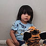 child, toddler, plush_toy, tiger, carpet, indoor, barefoot, blue_shirt, sitting, wall, toy, young_child, person, cute, casual_clothing, home, floor, hair, looking, stuffed_animal