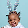 baby, smiling, bunny_ears, headband, curly_hair, child, portrait, cute, happy, white_clothing, blue_background, infant, face, person, studio_photo, joyful, playful, fashion_accessory, closeup, adorable