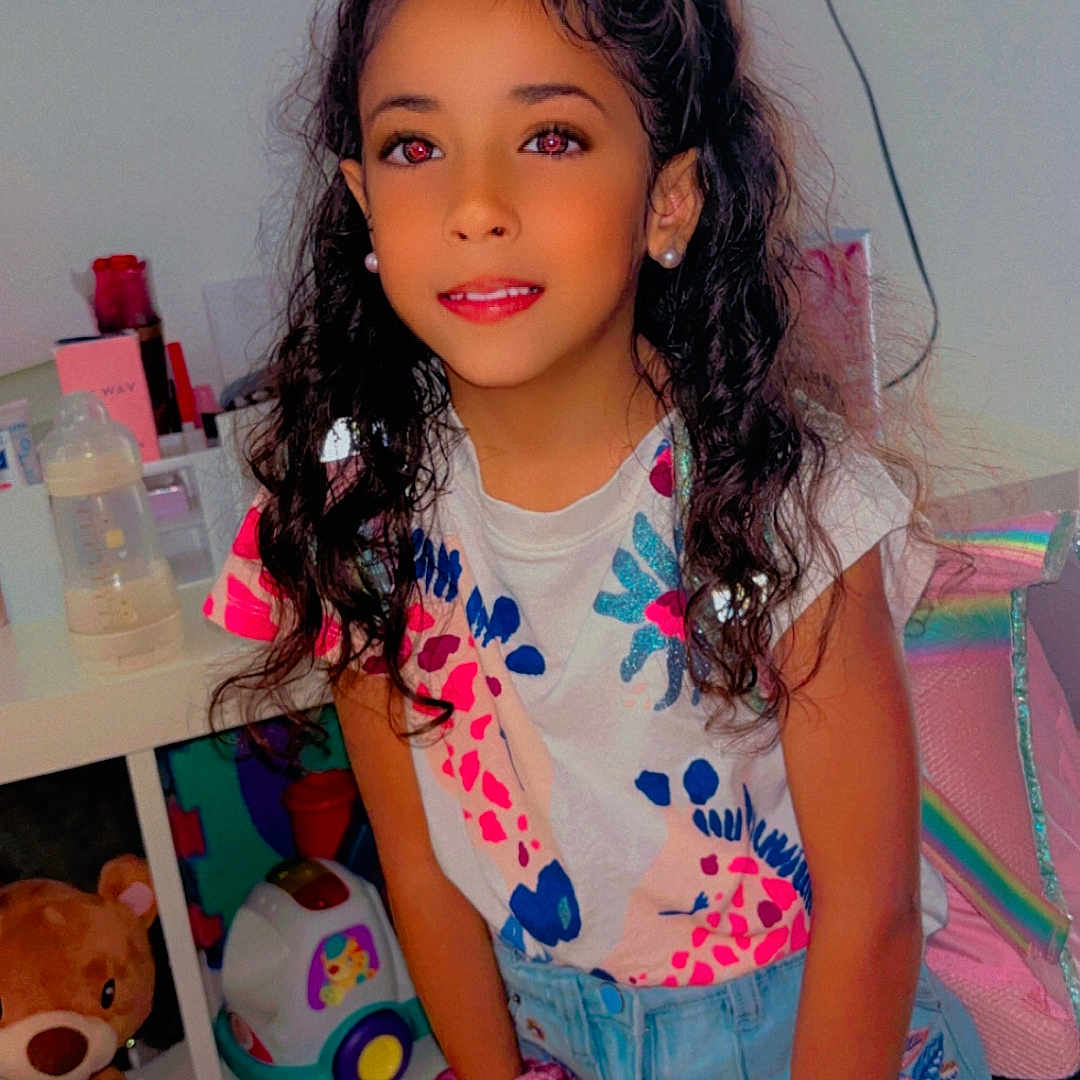 Djenna participe au concours pour gagner de l'argent avec cette photo : bottle, casual_clothing, child, childhood, colorful_shirt, curly_hair, denim_skirt, earrings, furniture, girl, hair_accessory, indoor, person, playful, portrait, room, smile, teddy_bear, toy, youth