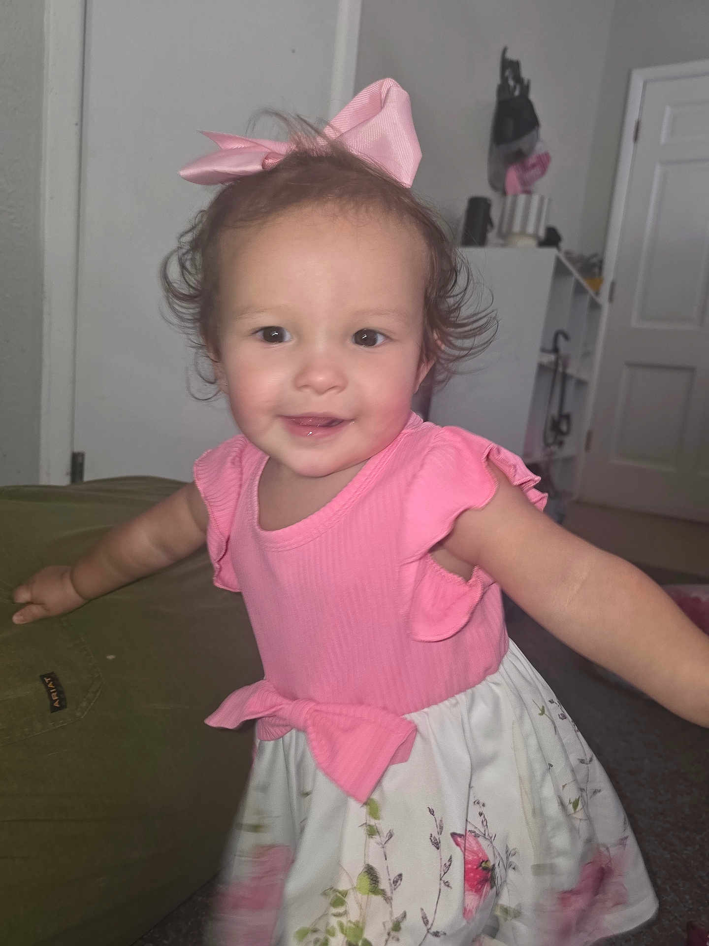 Jayla is registered to the contest to win money with this photo: toddler, child, pink_dress, bow, smiling, indoor, carpet, furniture, door, cabinet, curly_hair, happy, person, young_child, playful, cute, portrait, face, arms_outstretched, home