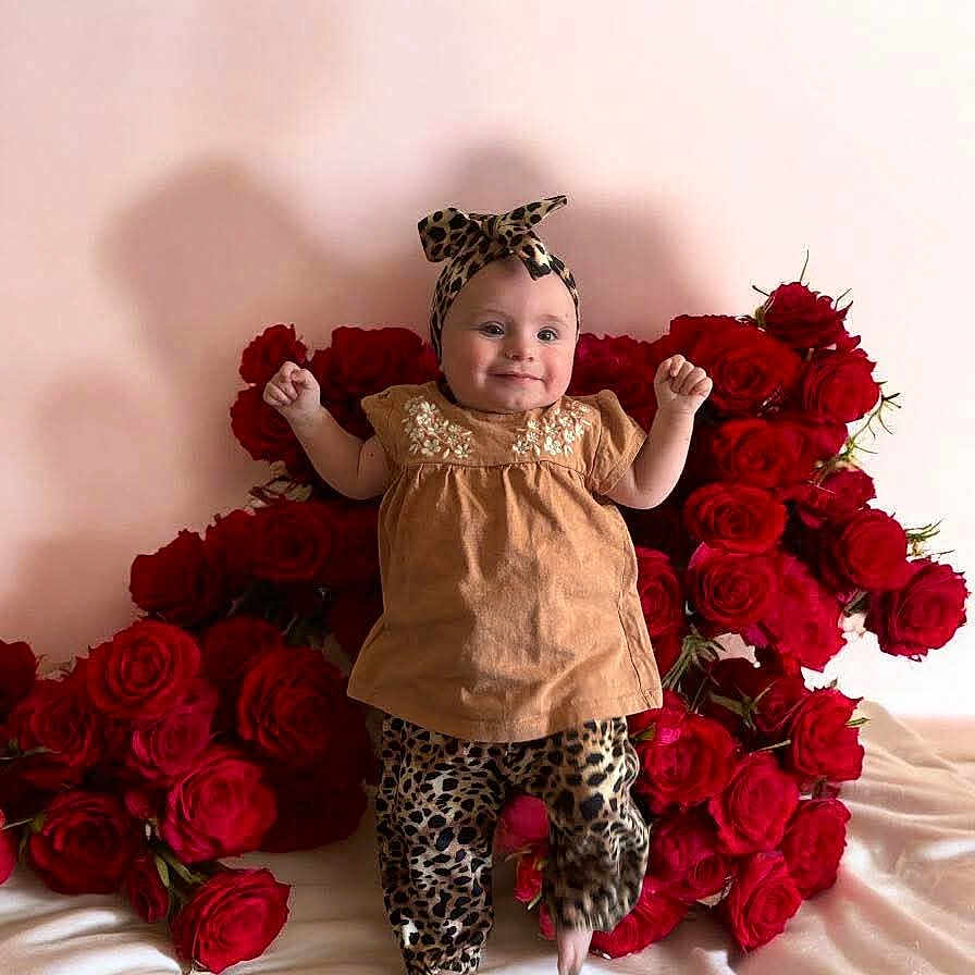 Deyva a rejoint le concours — aidez-le/la à gagner de superbes lots ! baby, brown_top, cheerful, child, cute, decor, floral, flower, happy, headband, indoors, infant, leopard_print, person, pink_background, portrait, red_roses, roses, sitting, smiling