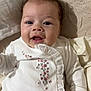 baby, infant, smiling, curly_hair, white_clothing, floral_embroidery, ruffles, blanket, soft_texture, closeup, portrait, cute, happy, child, indoor, face, hand, skin, young, cozy