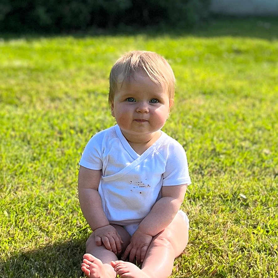Azélie a rejoint le concours — aidez-le/la à gagner de superbes lots ! baby, casual, child, cute, daylight, face, feet, grass, greenery, hands, happy, lawn, nature, onesie, outdoor, portrait, sitting, sunlight, white_clothing, young_child