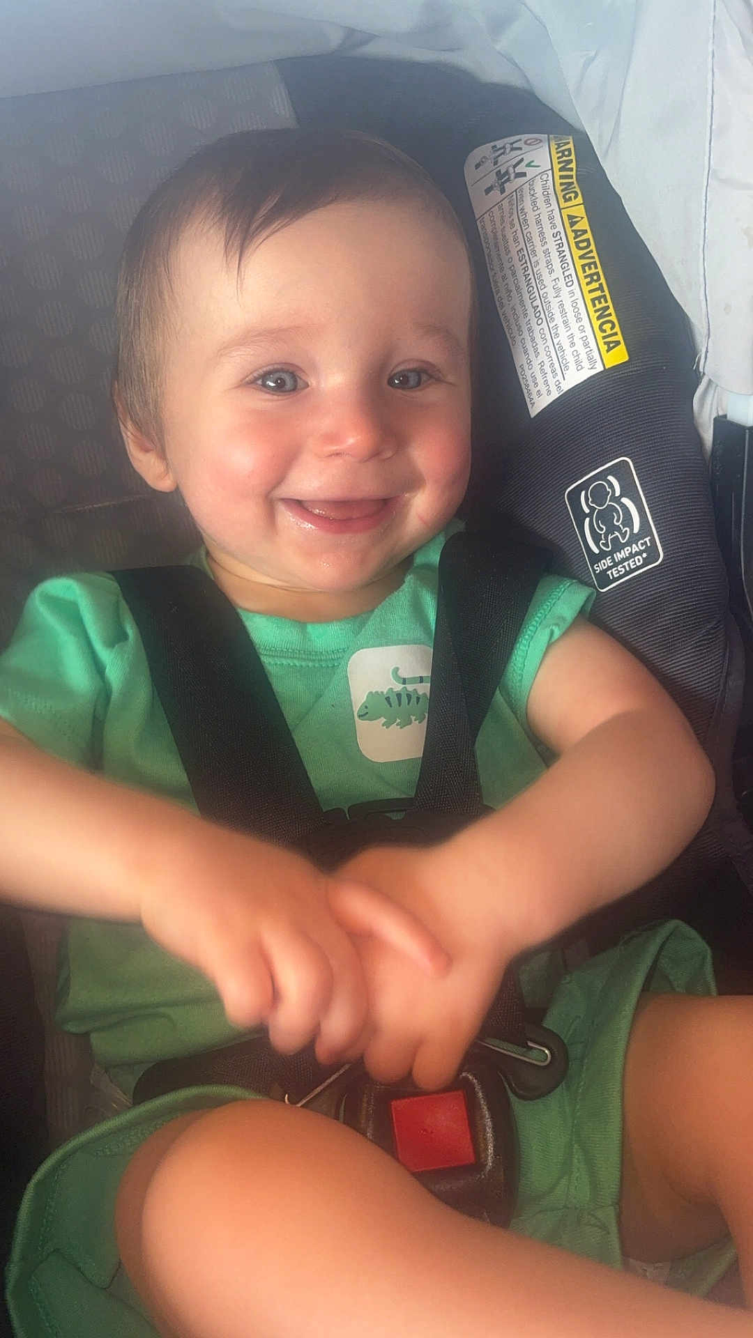 Liam is registered to the contest to win money with this photo: baby, child, smiling, car_seat, green_clothing, dinosaur_patch, straps, safety_harness, happy, cute, infant, person, seatbelt, indoors, hands, face, head, shoulder, legs, toddler