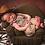 baby, newborn, sleeping, cowboy_hat, hat, overalls, knit_chair, crocheted_chair, props, little_feet, portrait, studio, cozy, brown, wooden_wheel, cowhide_rug, hands, face, skin, peaceful