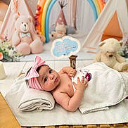 Diana is registered to the contest to win money with this photo: baby, infant, towel, pink_bow, headband, teddy_bear, rainbow, plush_toy, wooden_floor, blanket, cute, child, indoor, soft_lighting, portrait, laying_down, nursery, decor, toy, curious