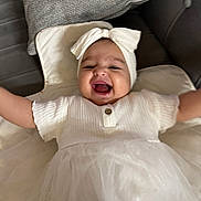 Diana is registered to the contest to win money with this photo: baby, child, smile, laughing, headband, white_dress, arms_outstretched, cushion, soft_surface, happy, infant, cute, indoor, portrait, toddler, headwear, button, fabric, clothing, person