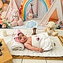 baby, infant, pink_bow, towel, wooden_mat, teddy_bear, rainbow, balloons, tent, stuffed_animal, nursery, soft_toys, flower_decoration, candle, blanket, headband, infant_face, indoor, cute, relaxing