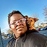 animal, blue_sky, car, casual, daytime, dog, glasses, happy, jacket, outdoor, person, pet, portrait, puppy, shoulder, smile, street, sunlight, tree, urban