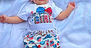 Omie is registered to the contest to win money with this photo: baby, child, smiling, headband, white_blanket, outdoor, grass, clothing, toddler, cute, summer, happy, person, portrait, infant, patterned_clothes, lying_down, celebration, festive, american_flag