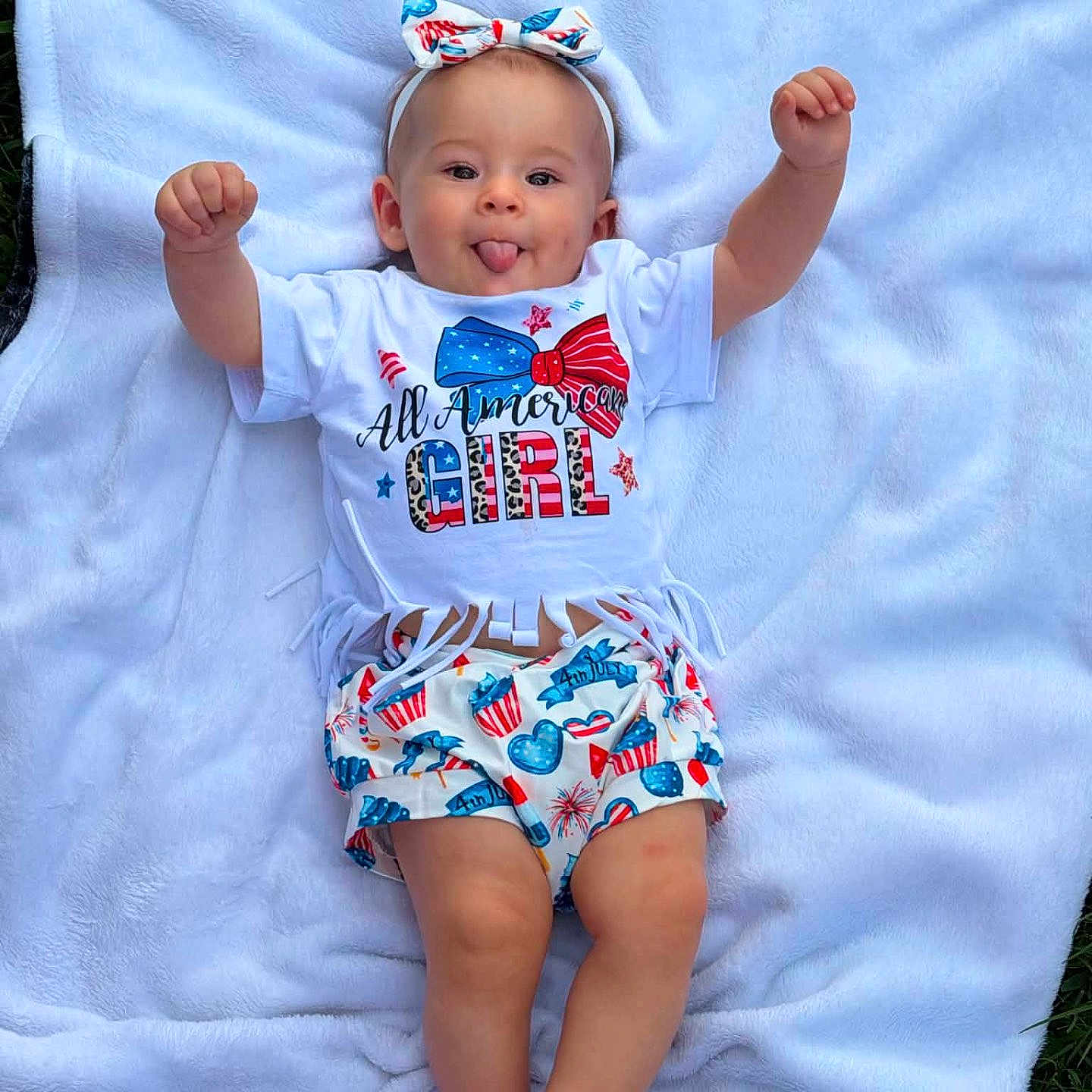 Omie joined the competition — help win amazing prizes! baby, child, tongue_out, headband, bow, white_blanket, outdoor, grass, shorts, tshirt, smiling, cute, celebration, 4th_of_july, red_white_blue, patterned_clothing, happy, playful, infant, person