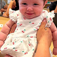 Charlie is registered to the contest to win money with this photo: baby, infant, child, smiling, portrait, red_bow, dress, floral_pattern, sitting, held_baby, adult_hand, indoor, store, shop, wooden_counter, background_people, face, happy, fashion, photography