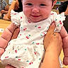 baby, infant, child, smiling, portrait, red_bow, dress, floral_pattern, sitting, held_baby, adult_hand, indoor, store, shop, wooden_counter, background_people, face, happy, fashion, photography
