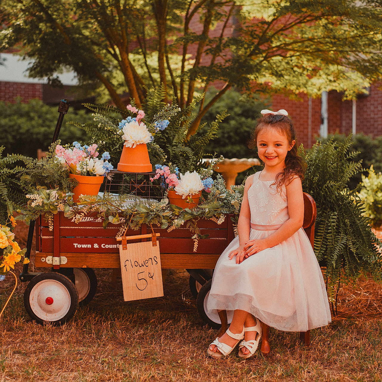Paisley joined the competition — help win amazing prizes! child, cute, flower_pots, flowers, garden, girl, grass, greenery, handwritten_sign, nature, outdoor, plants, sign, smile, spring, sunlight, trees, wagon, white_dress, wooden_chair