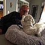 man, dog, pet_bed, indoor, glasses, white_dog, fluffy_bed, interaction, affection, living_room, curtains, furniture, television, wall_art, casual_clothing, home, paw, light, window, relaxation