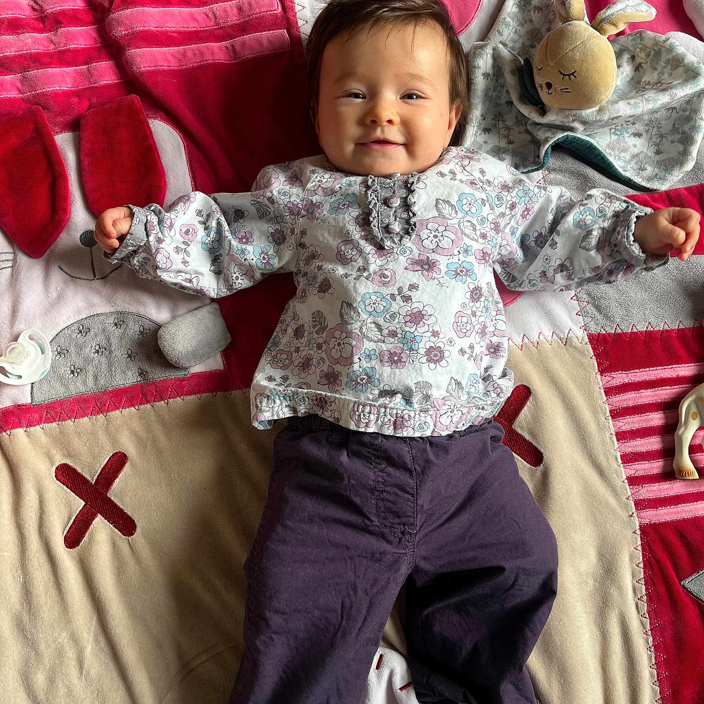 Julia participe au concours pour gagner de l'argent avec cette photo : baby, blanket, casual_clothing, child, colorful, cute, face, floor, happy, head, indoor, infant, lying_down, pacifier, patterned_clothing, person, playtime, plush_toy, smiling, soft_toy