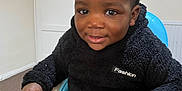 Aadil is registered to the contest to win money with this photo: toddler, child, smiling, indoor, black_clothing, sweater, chair, curly_hair, happy, person, seated, fashion_label, carpet, door, radiator, hand, face, portrait, young_child, cozy