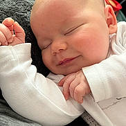 Logan is registered to the contest to win money with this photo: baby, sleeping, smile, infant, white_clothing, hand, face, peaceful, closeup, soft_texture, skin, cozy, resting, cute, child, portrait, newborn, relaxation, indoors, warm_light
