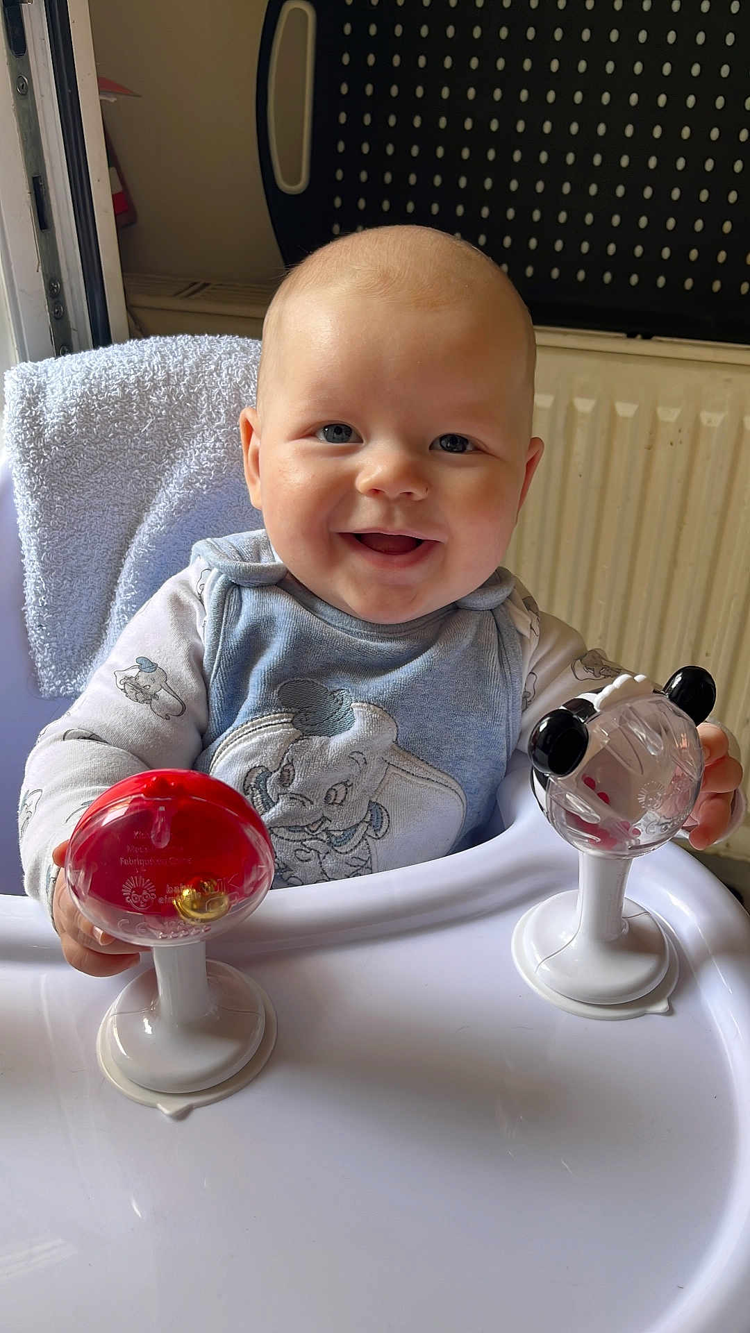 Logan is registered to the contest to win money with this photo: baby, child, high_chair, toy, smiling, indoor, towel, radiator, clothing, face, person, happy, holding, sitting, cute, infant, playtime, closeup, head, blue_eyes