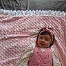 adorable, baby, bed_sheet, blanket, bow, cozy, dark_hair, embroidered_text, headband, infant, lying_down, pink_blanket, pink_outfit, plush_texture, portrait, ruffle_edge, small_hands, smiling, sunlight, white_shirt