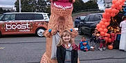 Julianne is registered to the contest to win money with this photo: child, costume, inflatable, t_rex, dinosaur, outdoor, parking_lot, halloween, pumpkins, balloons, kids, smile, candy_bucket, event, vehicle, trees, cloudy_sky, people, festive, celebration