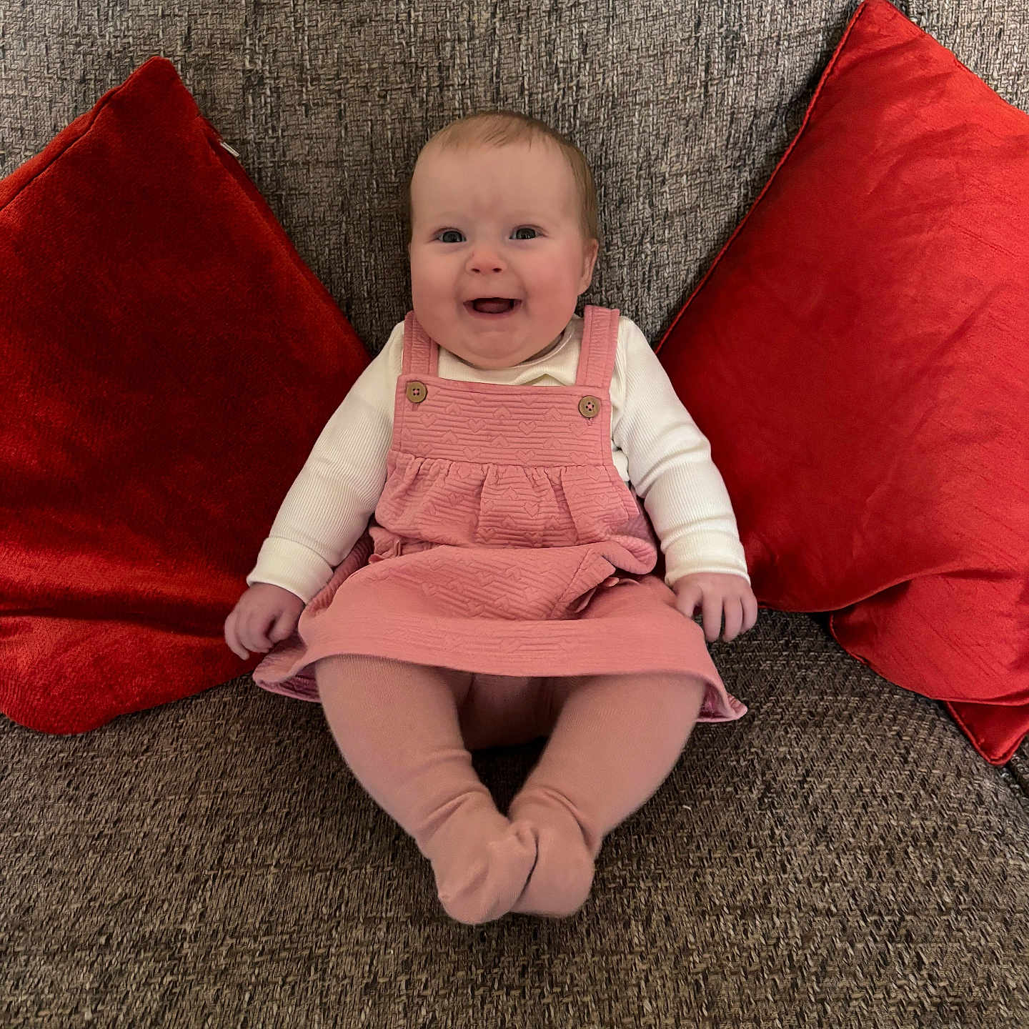 Mabel-Ivy is registered to the contest to win money with this photo: baby, child, comfort, couch, cozy, cute, furniture, happy, home, indoor, infant, living_room, person, pink_dress, portrait, red_cushions, seated, smiling, textile, tights