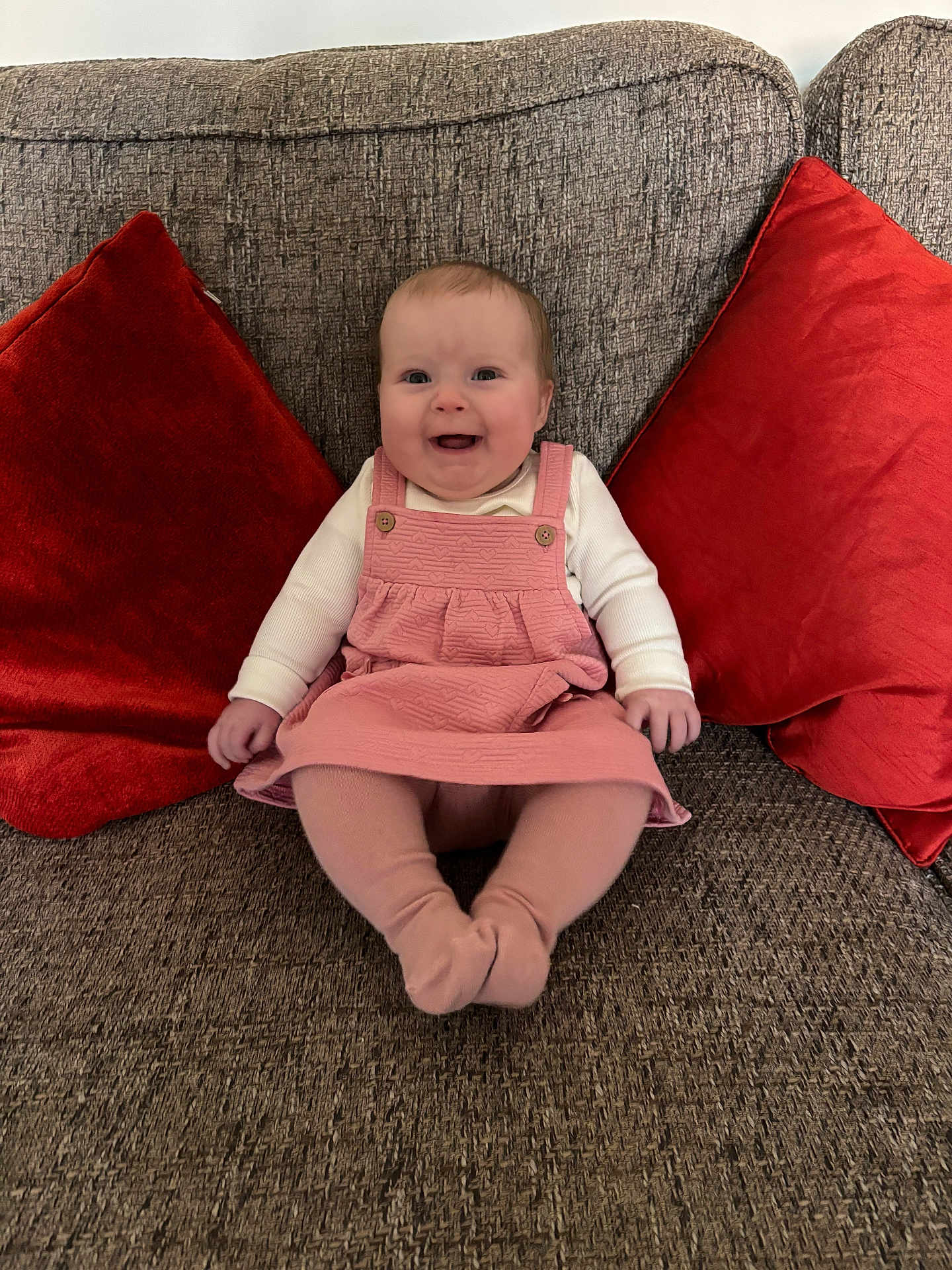 Mabel-Ivy is registered to the contest to win money with this photo: baby, child, smiling, pink_dress, tights, couch, red_cushions, indoor, portrait, happy, seated, infant, home, furniture, cute, person, cozy, living_room, textile, comfort