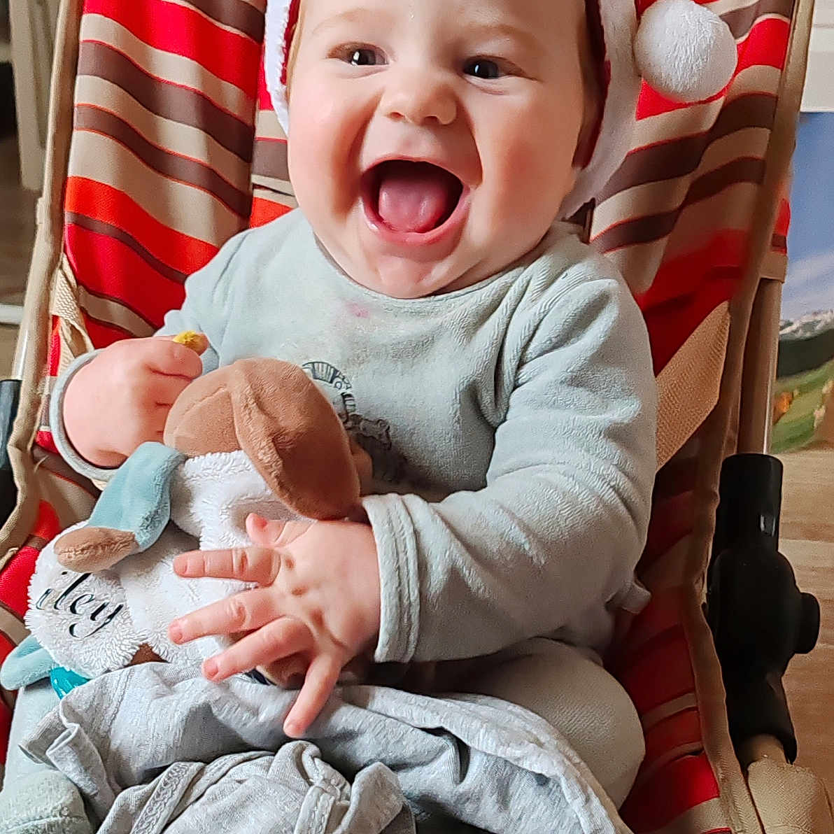 Riley a rejoint le concours — aidez-le/la à gagner de superbes lots ! baby, blanket, child, clothing, cute, face, hands, happy, holiday, indoor, infant, joyful, person, plush_toy, red, santa_hat, seated, smiling, striped_chair, white