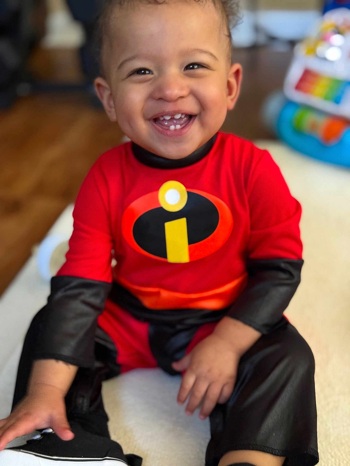 Joseph joined the competition — help win amazing prizes! toddler, child, smiling, happy, superhero_costume, red_clothing, black_clothing, indoor, toy, blurred_background, sitting, portrait, cute, baby, person, face, hand, footwear, wooden_floor, home
