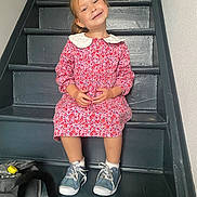 Lola participe au concours pour gagner de l'argent avec cette photo : child, girl, smiling, stairs, red_dress, white_collar, blue_sneakers, white_socks, missing_tooth, black_cat_tail, backpack, indoor, portrait, happy, casual, seated, wooden_stairs, playful, cute, young