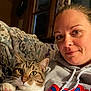 woman, cat, tabby_cat, indoor, hoodie, couch, blanket, window, curtain, pet, face, portrait, cozy, relaxed, whiskers, paw, feline, human, casual, smile
