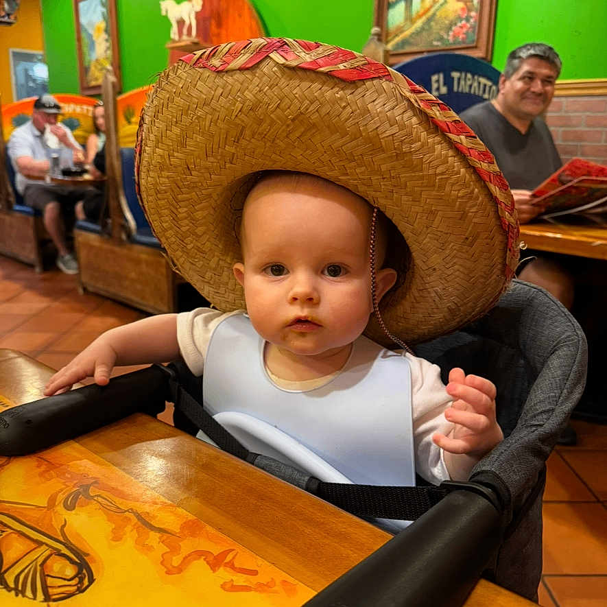 James is registered to the contest to win money with this photo: baby, casual, child, colorful, curious, dining, family, green_wall, hat, high_chair, indoor, infant, people, person, restaurant, seated, sombrero, table, wall_art, wooden_table