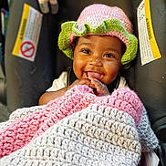 Apple is registered to the contest to win money with this photo: accessory, baby, blanket, car_seat, close_up, crocheted_blanket, crocheted_hat, cute, hands, happy, hat, indoor, infant, portrait, seat_harness, smile, smiling, tongue, toy, warm_lighting