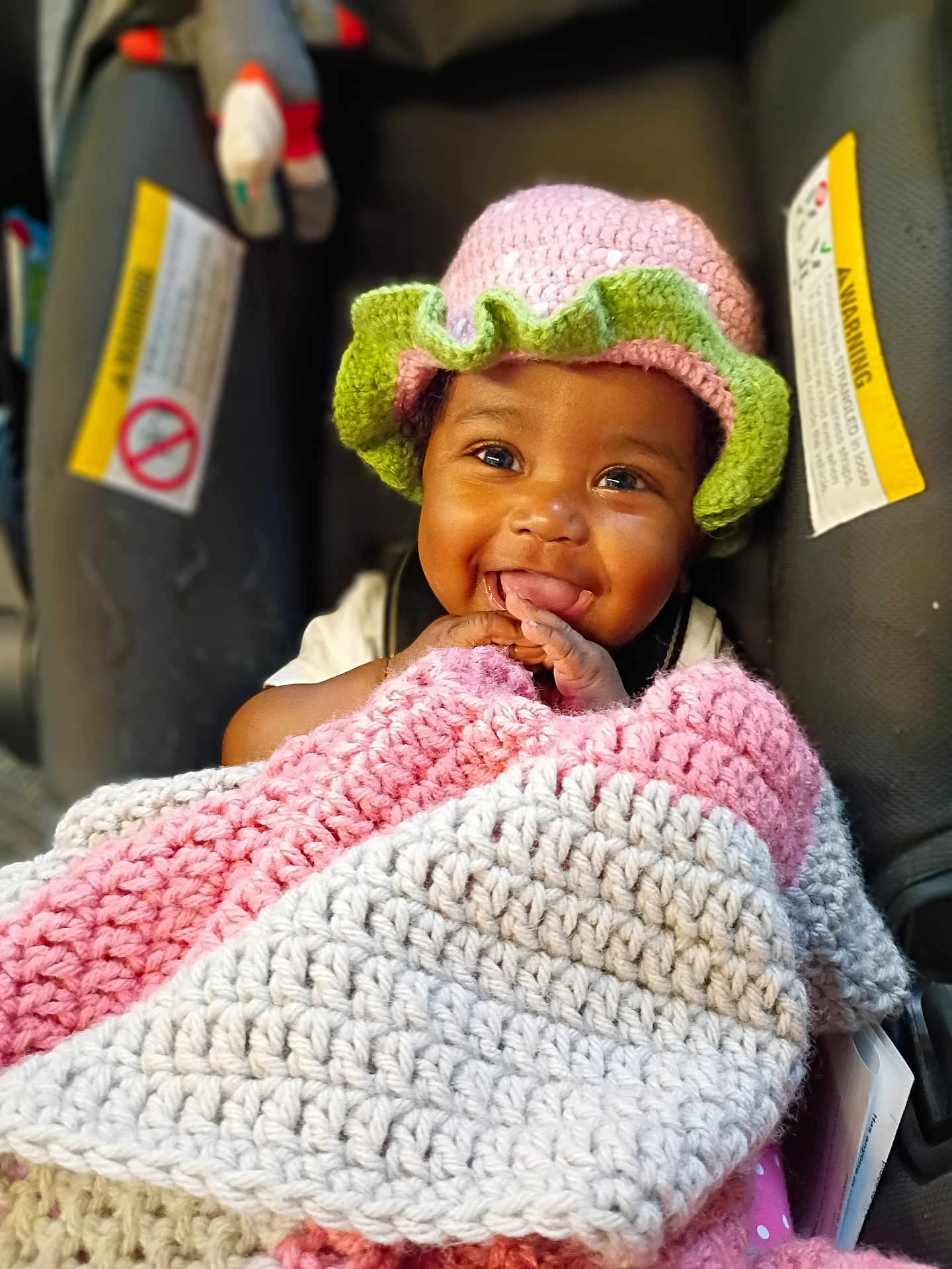 Apple is registered to the contest to win money with this photo: accessory, baby, blanket, car_seat, close_up, crocheted_blanket, crocheted_hat, cute, hands, happy, hat, indoor, infant, portrait, seat_harness, smile, smiling, tongue, toy, warm_lighting