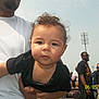 baby, child, person, curly_hair, outdoor, daylight, adult, crowd, stadium_lights, black_shirt, white_shirt, hand, face, head, skin, people, event, photography, portrait, casual