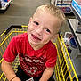 child, boy, shopping_cart, red_shirt, smiling, blue_eyes, toy_package, store_aisle, shopping, yellow_cart, shorts, casual_clothing, happy, portrait, retail, floor, shelves, product_display, indoor, cute