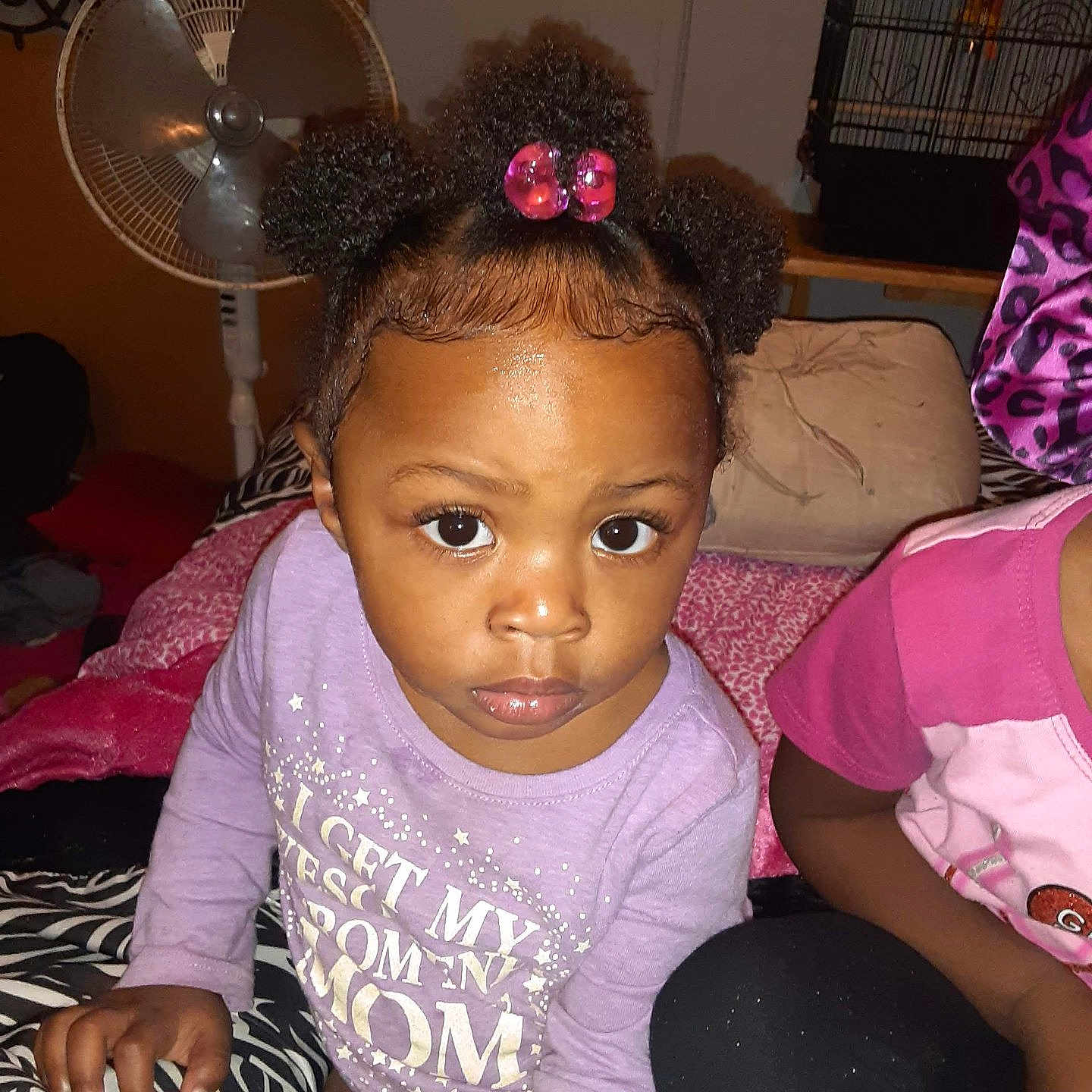 Za'Mierra Grace is registered to the contest to win money with this photo: bed, birdcage, black_pants, child, clothing, curious, eyes, face, fan, furniture, hair_puffs, hand, indoor, patterned_bedspread, person, phone, pink, purple_shirt, sitting, toddler